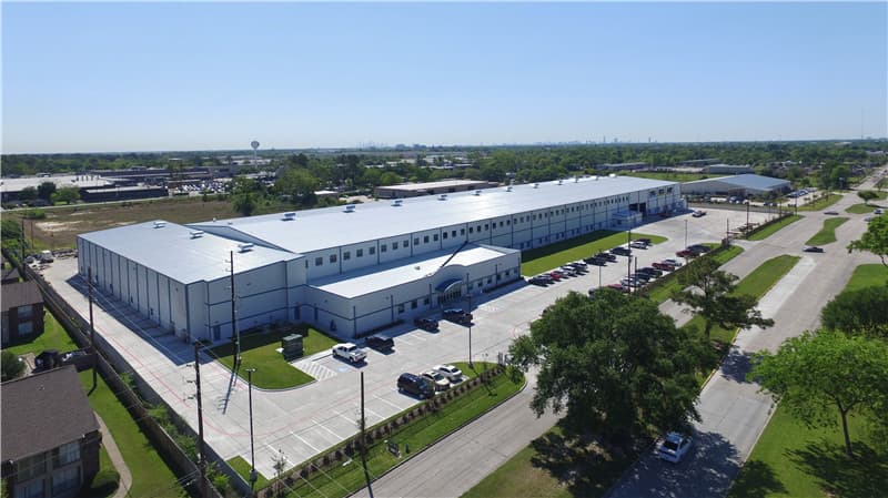 Aerial view of a large white industrial warehouse with a parking lot and surrounding greenery.